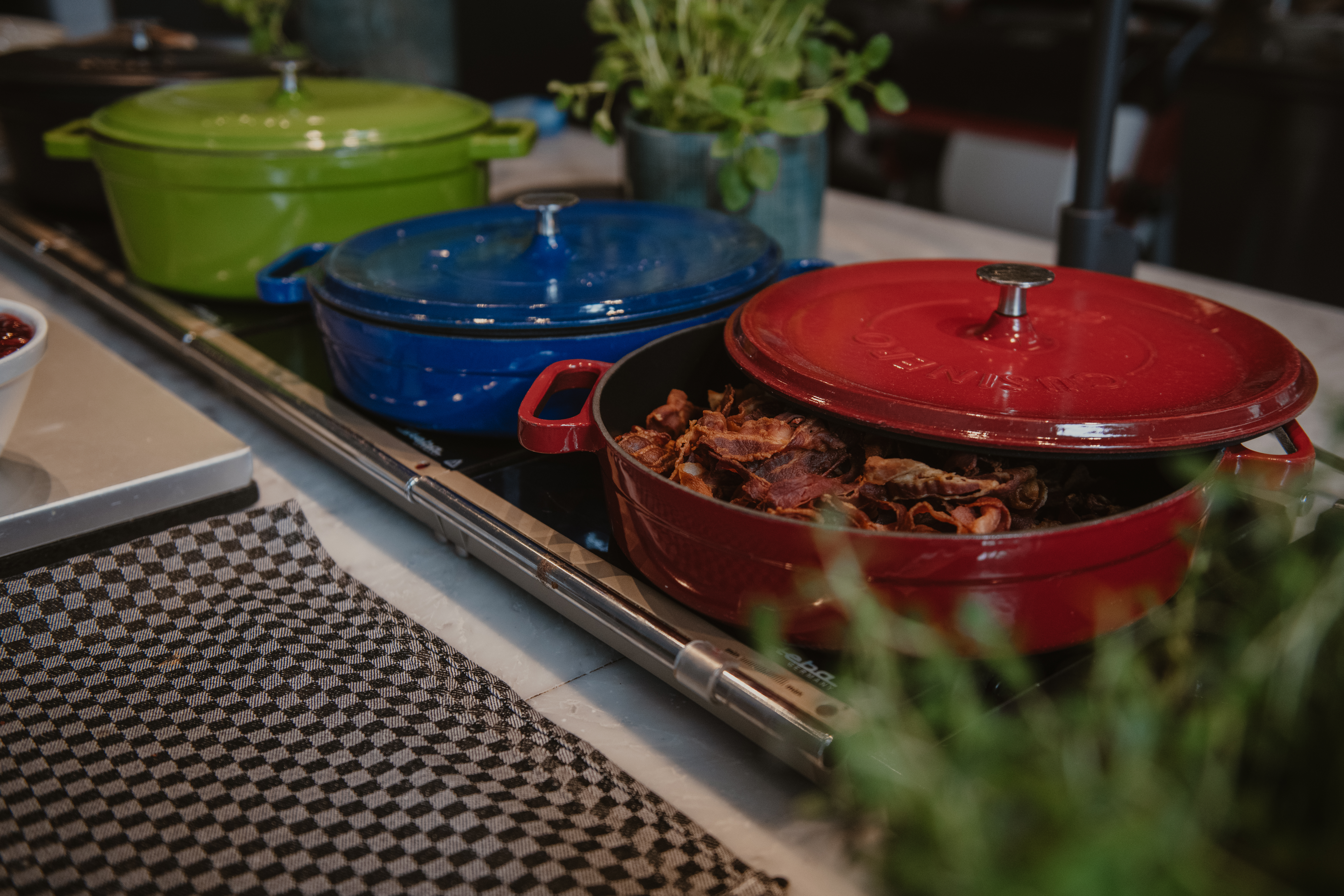 Röd gryta fylld med tillagat kött, delvis öppen på en bänk. Två grytor, en grön och en blå, syns i bakgrunden. Rutig duk och örter omger dem.