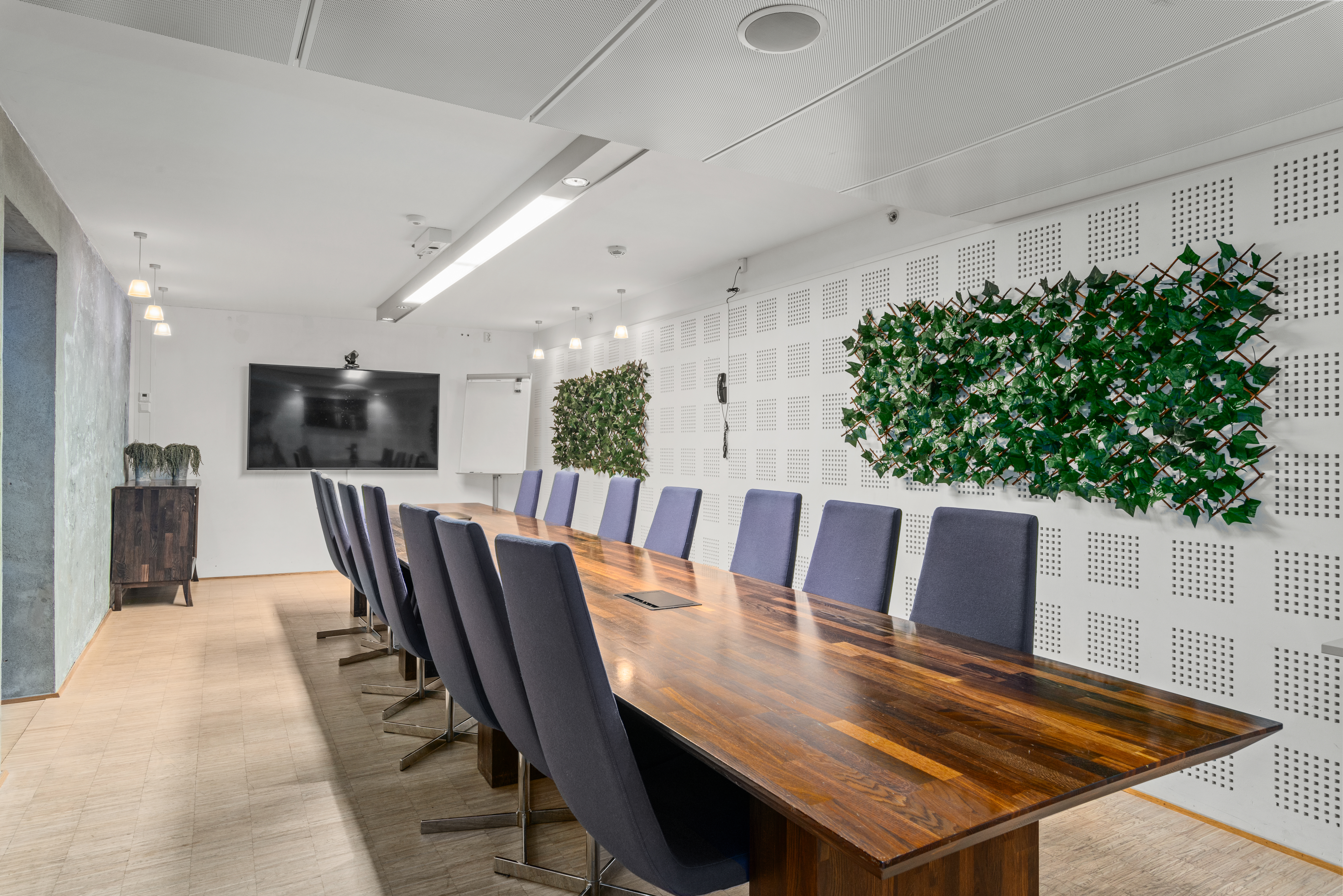 Et langt konferansebord i tre omgis av stoler i et moderne møterom med en veggmontert TV og dekorative planter på veggene.