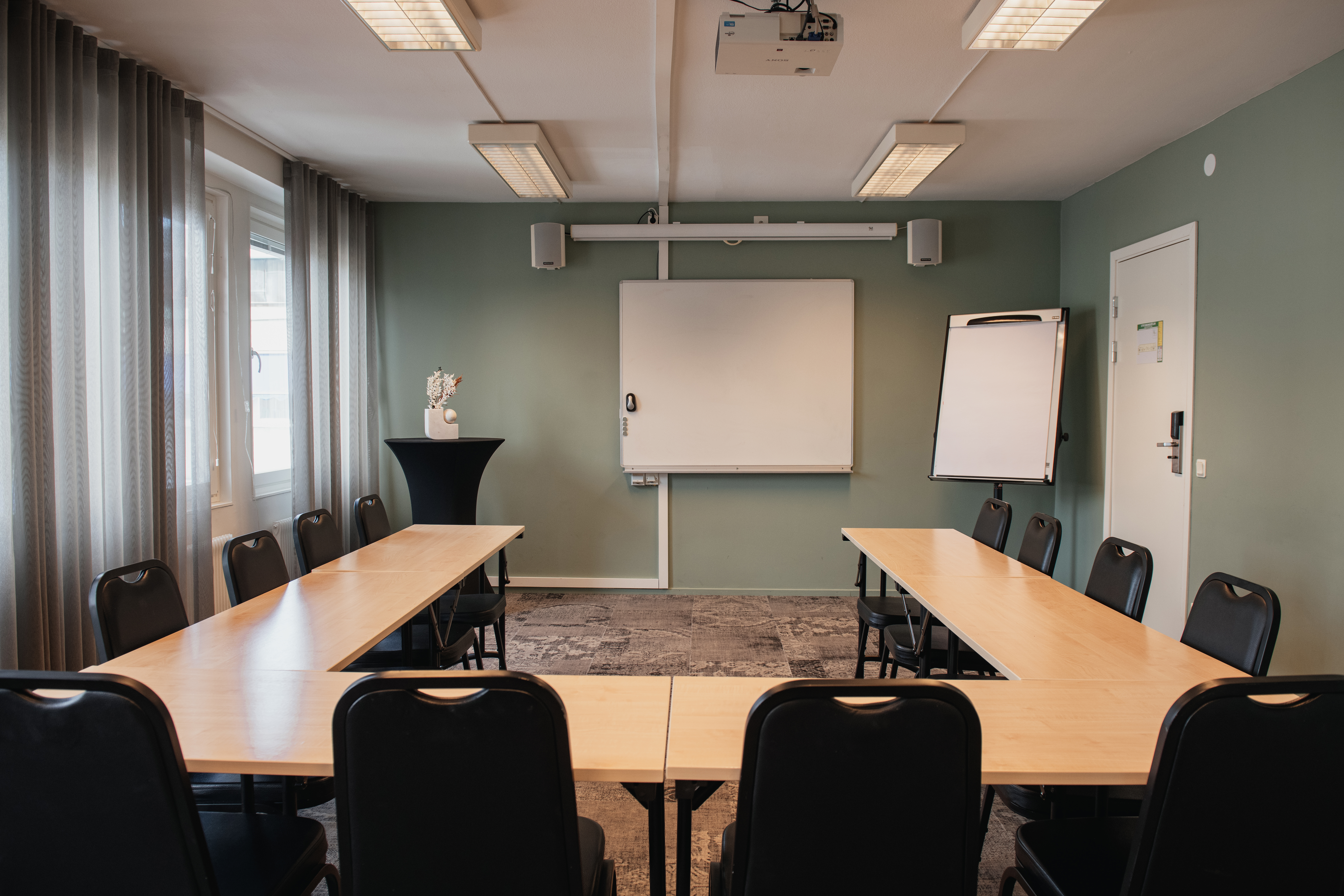 Konferencelokale med U-formede borde og stole. Et whiteboard og flipover står mod en grøn væg. Overheadlys oplyser rummet.