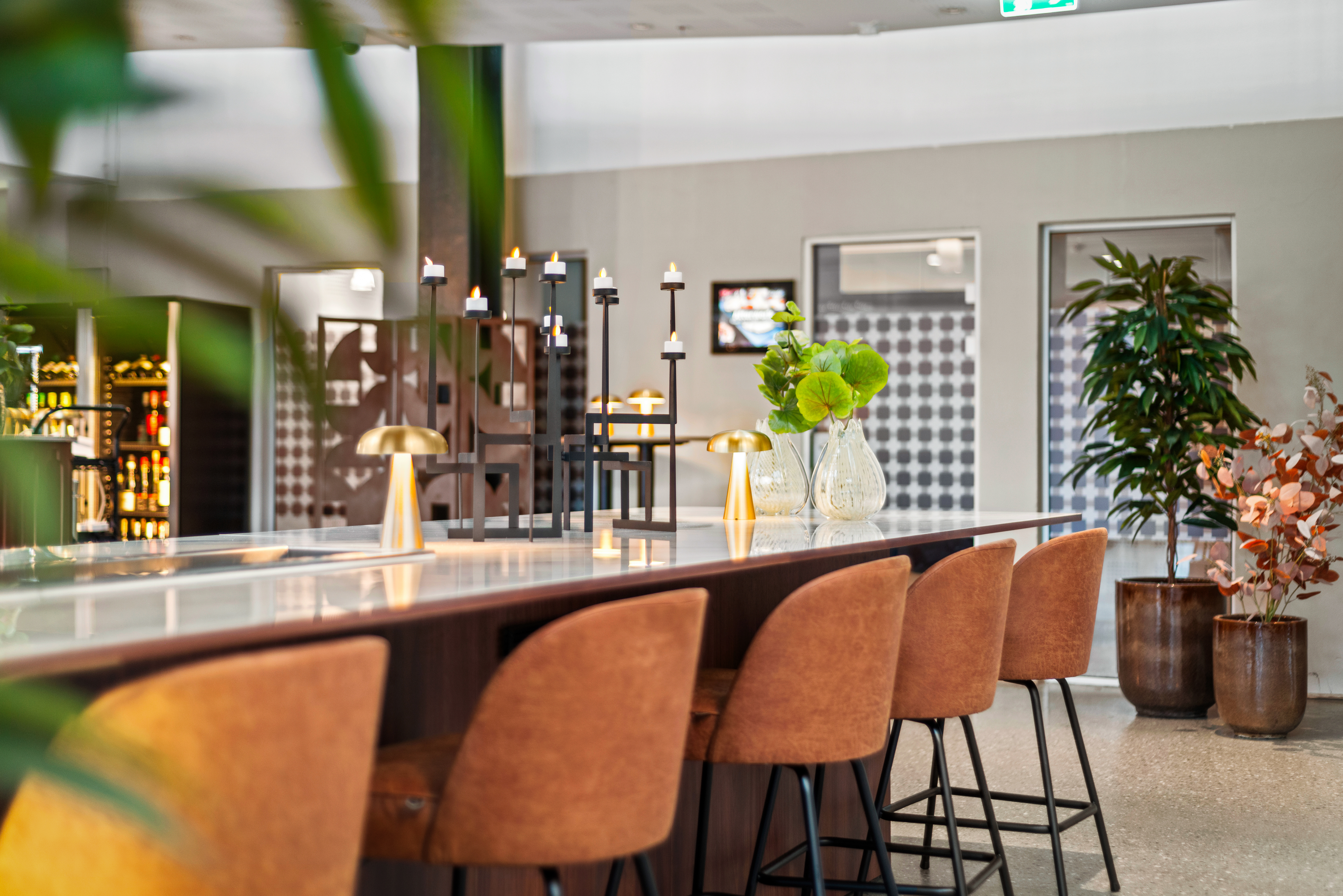 En lang, hvit benkeplate med brune stoler er pyntet med gullfargede lamper og vaser med grønne planter. Den står i et moderne, åpent rom med dekorative planter og hyller.