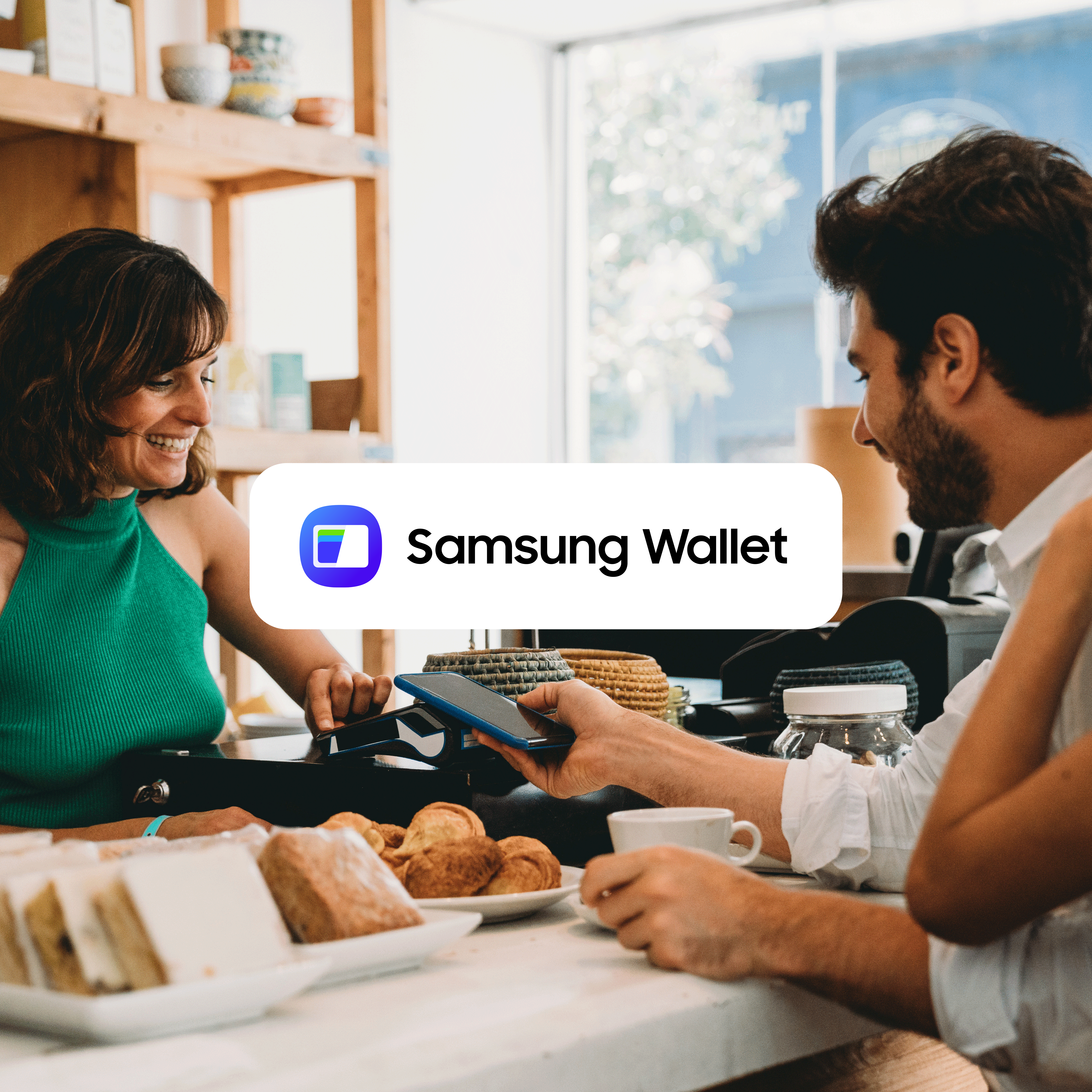Una mujer rubia, con un cárdigan verde y una blusa rosa, está sentada en una mesa de café y realiza un pago sin contacto con Samsung Wallet en su smartphone.